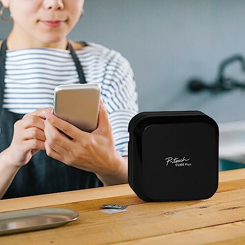 Person using smartphone near a black printer device on a table.